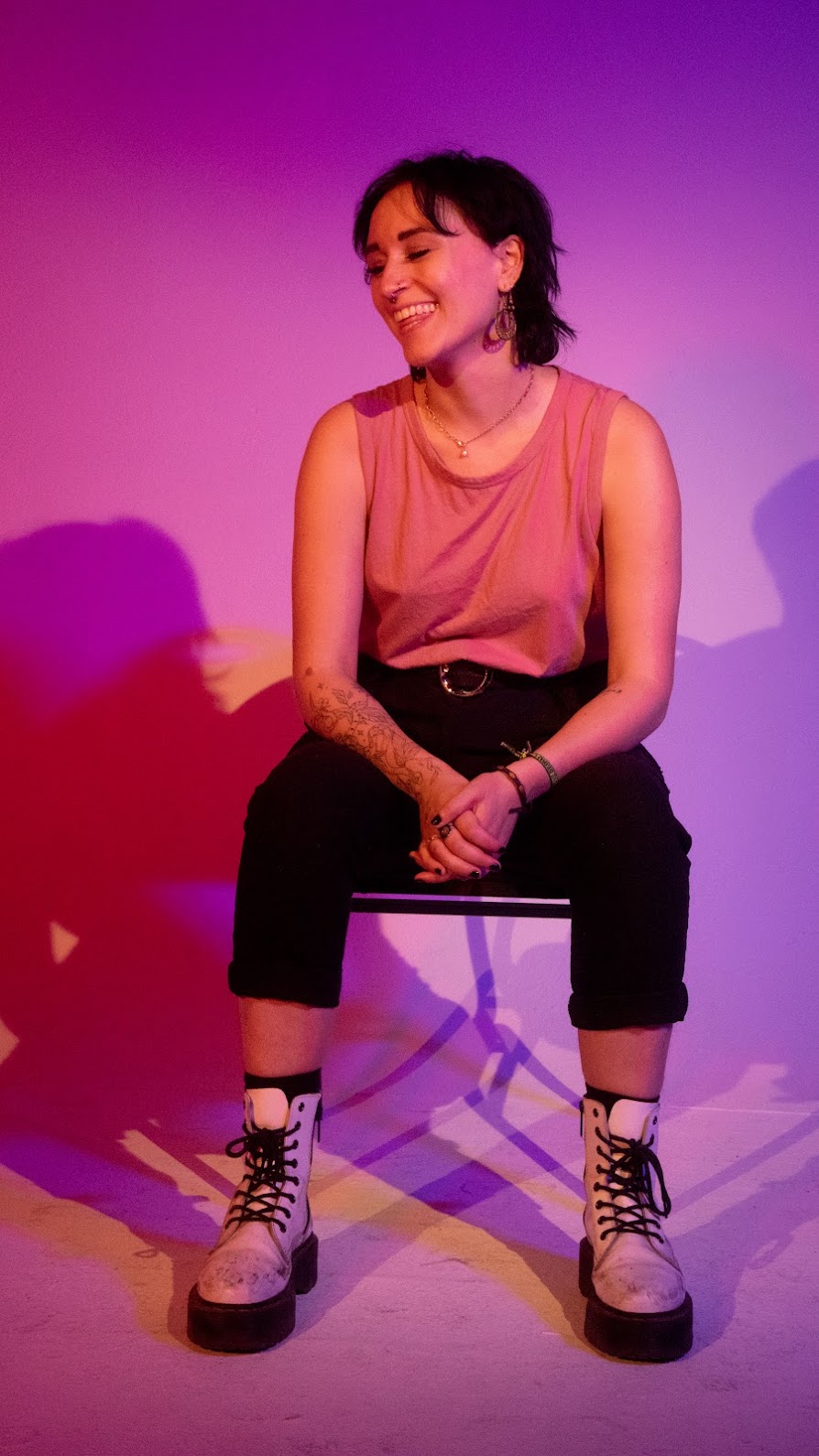 Tess, a Caucasian woman, has short black hair and is sitting in a studio setting with pink and purple lights behind her.