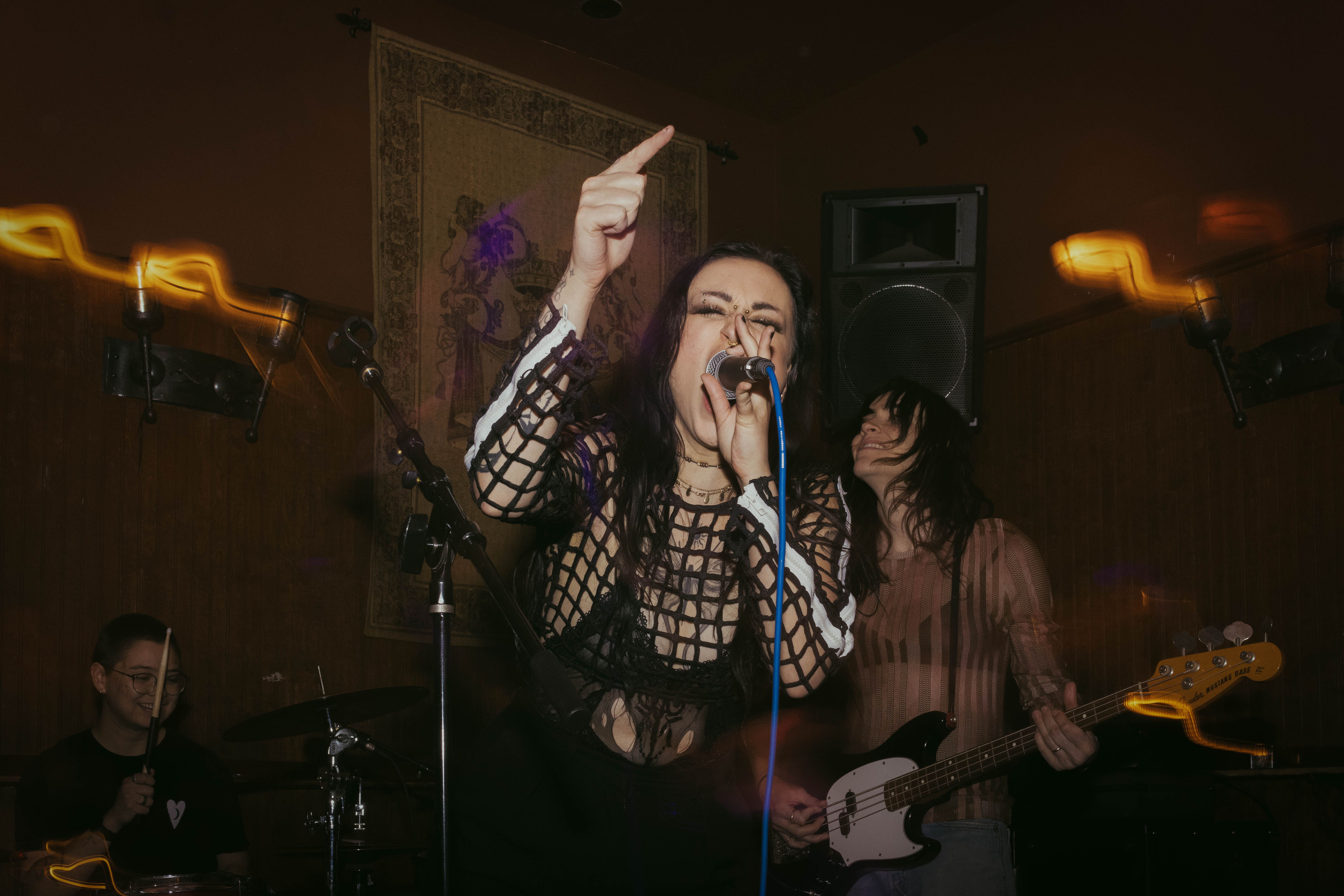 Tess, a Caucasian woman, singing into a microphone. Her hand is up with a finger pointed upwards, and she is singing loudly.