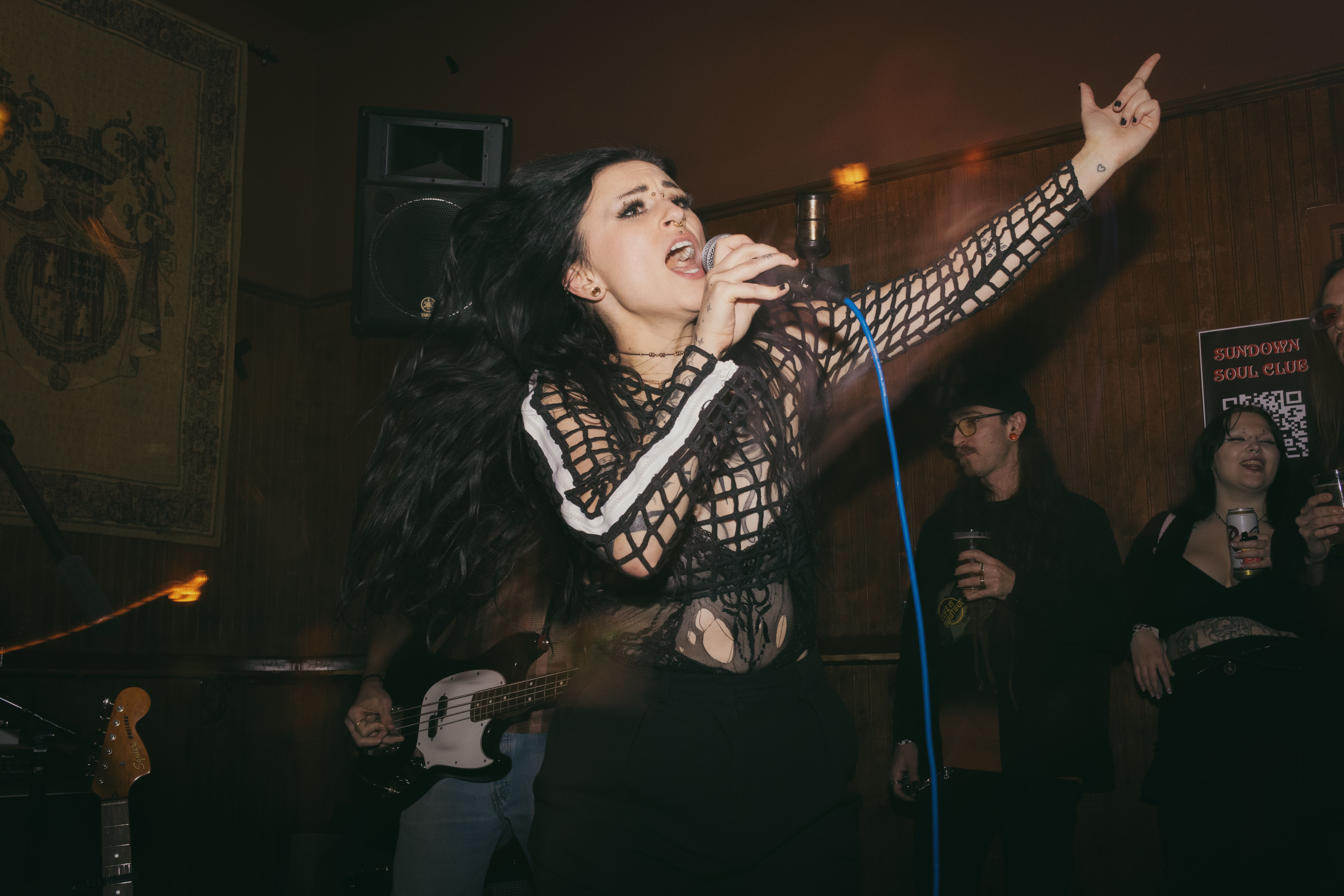 Tess, a Caucasian woman, singing into a microphone. Her hand is up and she is singing loudly.