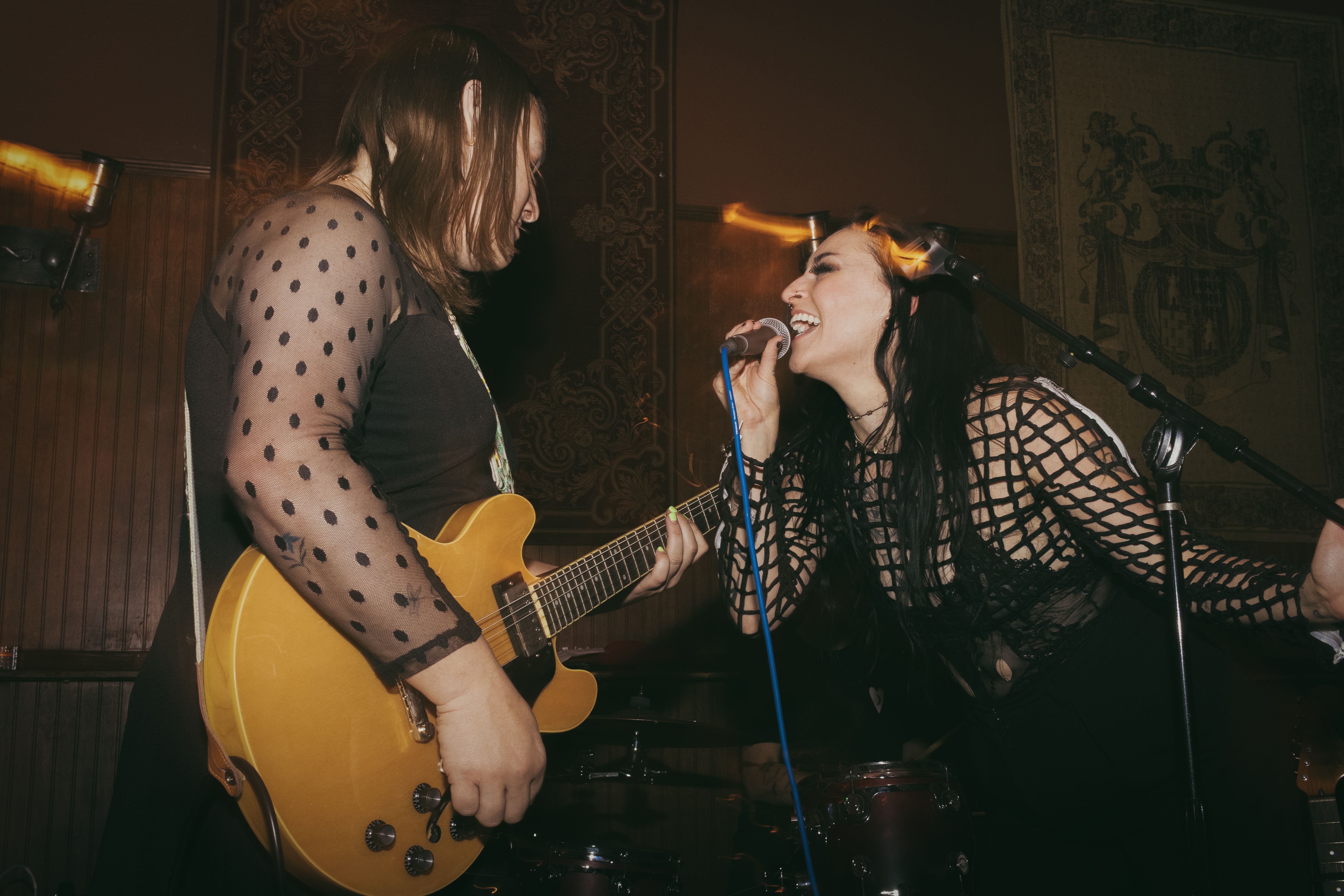 Tess, a Caucasian woman, singing facing a guitarist, another Caucasian woman with brown hair. The woman is playing a yellow guitar.
