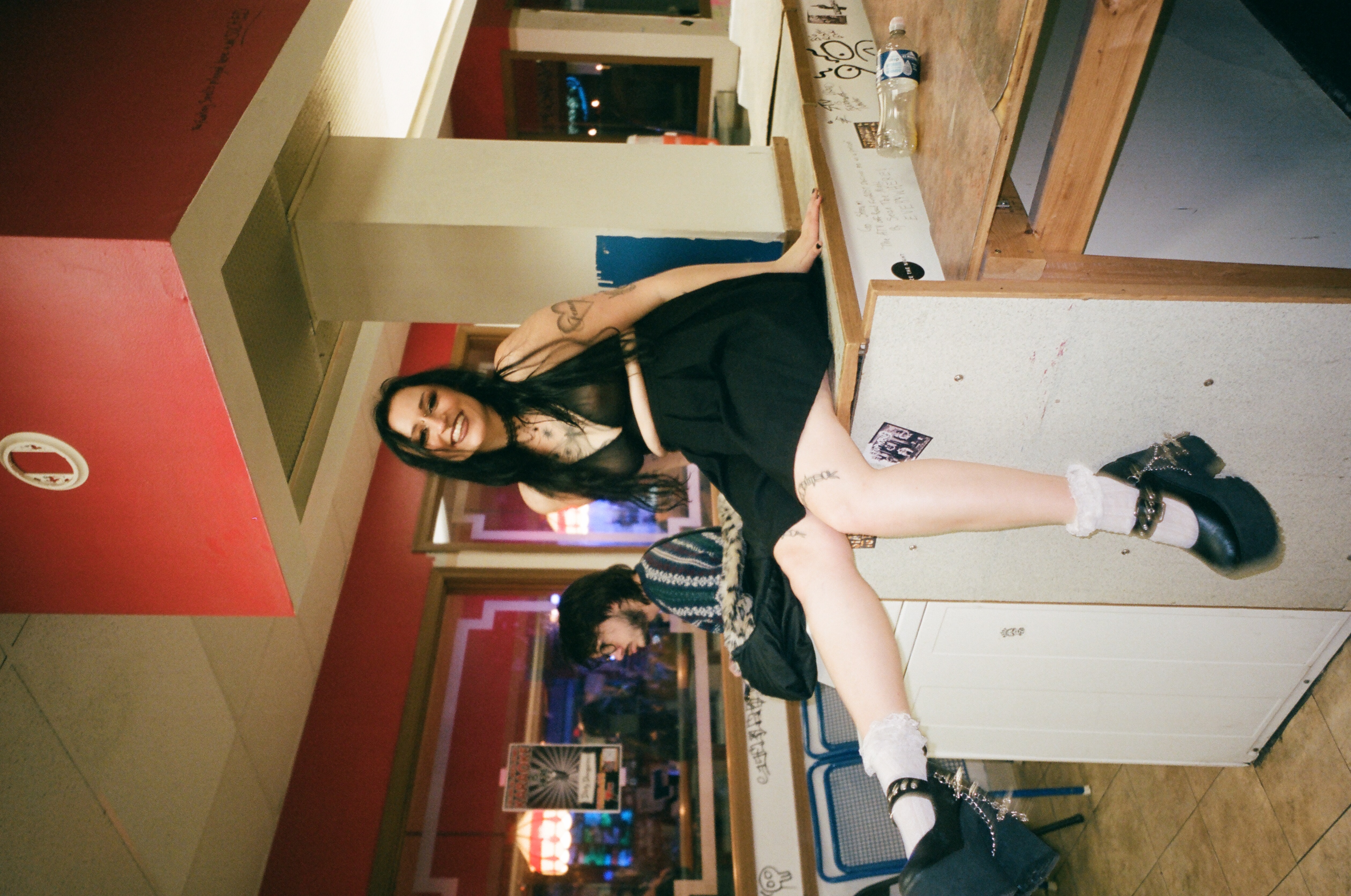 Tess, a Caucasian woman with long black hair and gold facial piercings, sits on a washing machine. She has some black tattoos. She is wearing a black skirt with a see-through black bra and chunky black heels. She is smiling at the camera. The photo has a sepia tone and is a bit grainy.