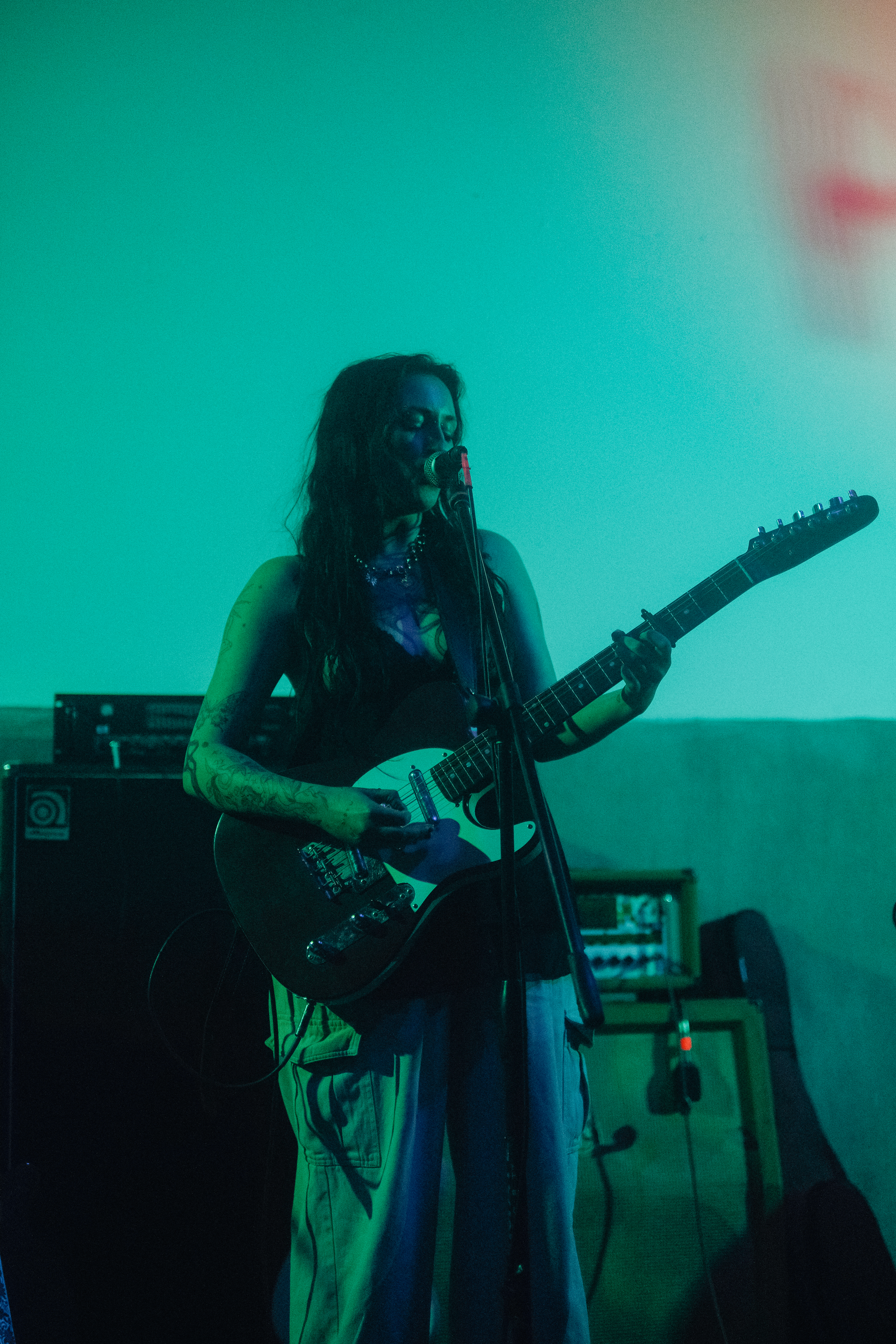 Tess, a Caucasian woman, is playing a red guitar on a stage with teal lighting. She is wearing tan pants and a black babydoll lingerie shirt.
