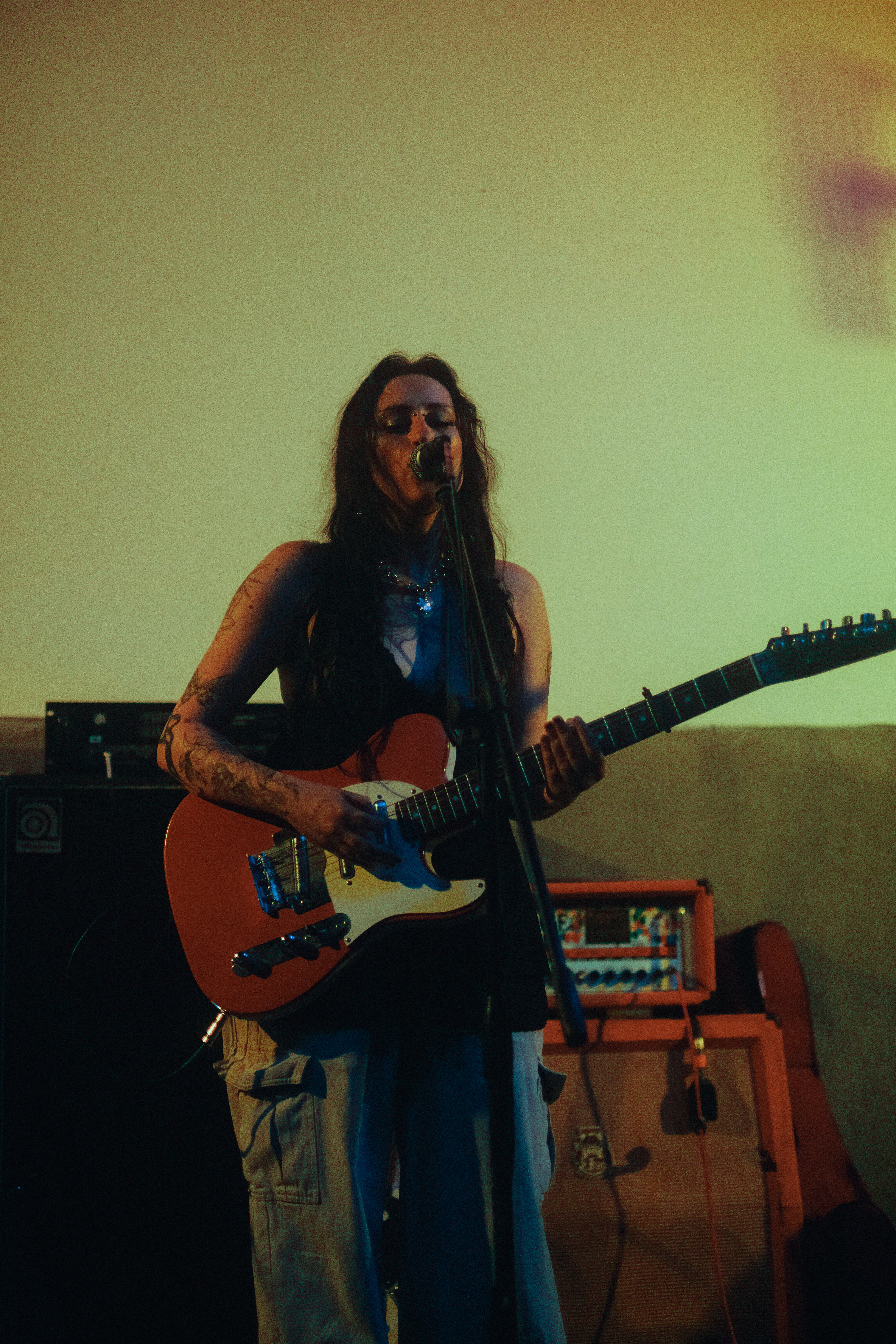 Tess, a Caucasian woman, is playing a red guitar on a stage with yellow lighting. She is wearing tan pants and a black babydoll lingerie shirt.