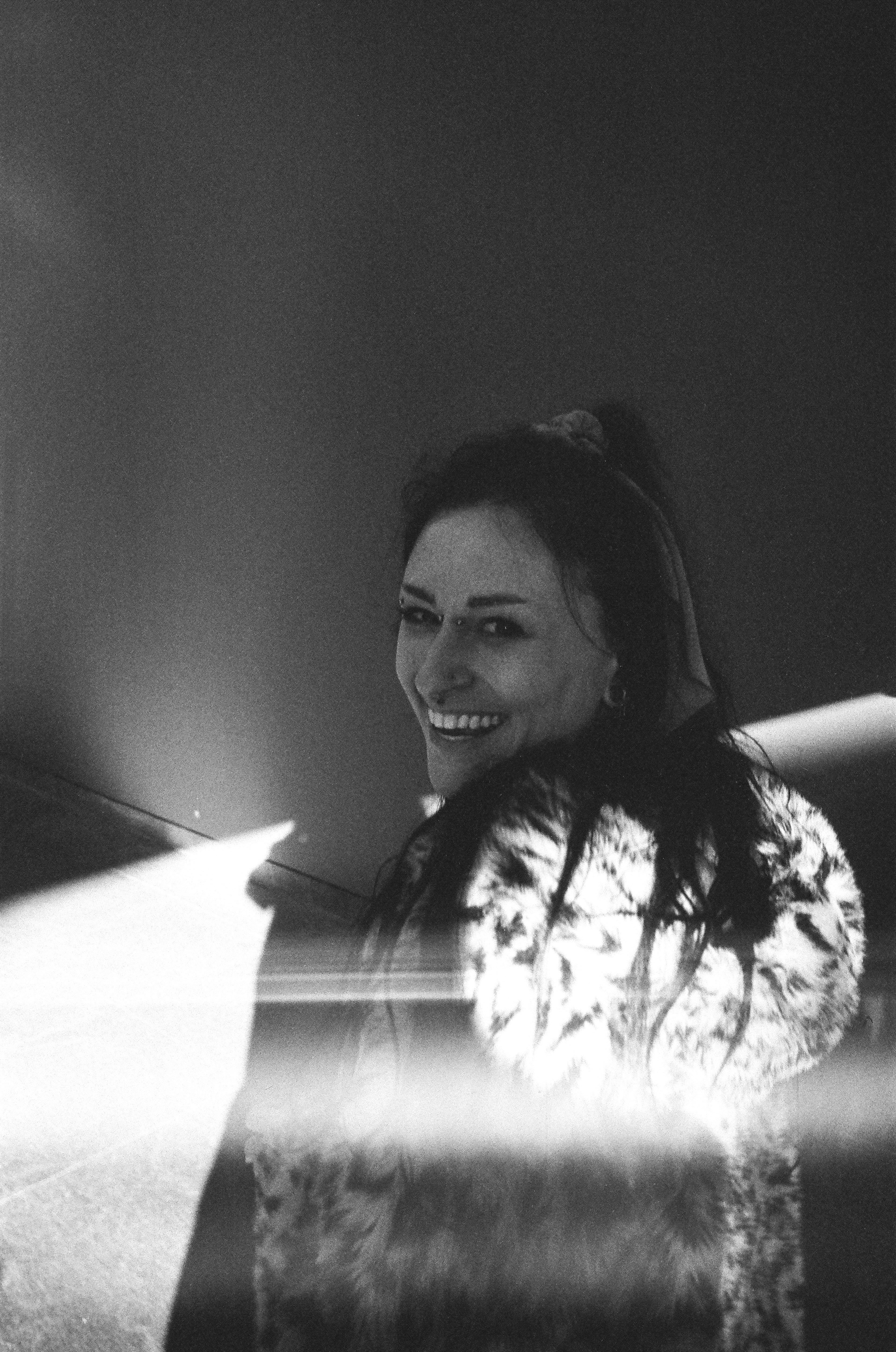 A black and white photo of Tess, looking back at the camera and smiling. She is wearing a fuzzy cheetah print coat.
